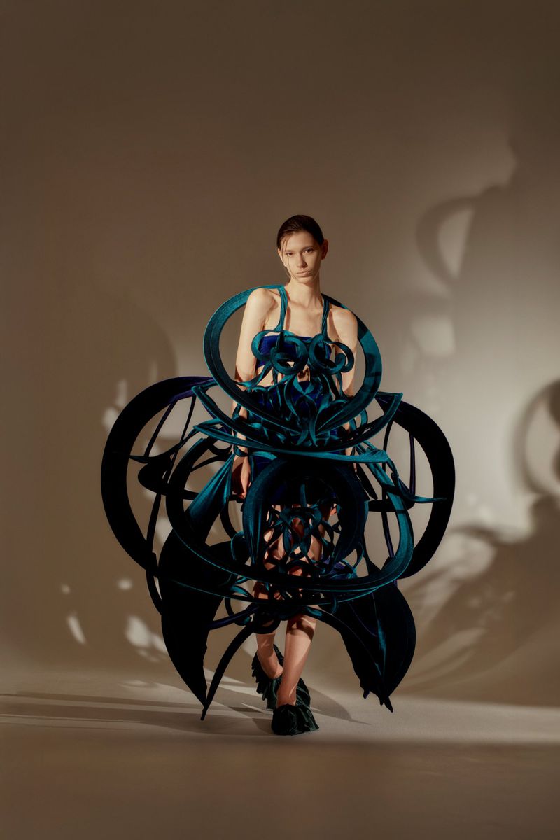 A full-length studio shot captures a model in an avant-garde blue dress with sculptural details, set against a neutral backdrop.