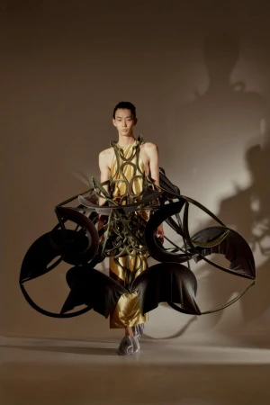Woman walks forward in a gold gown featuring circular structure surrounding the dress. Backlit, indoor setting with a neutral backdrop.
