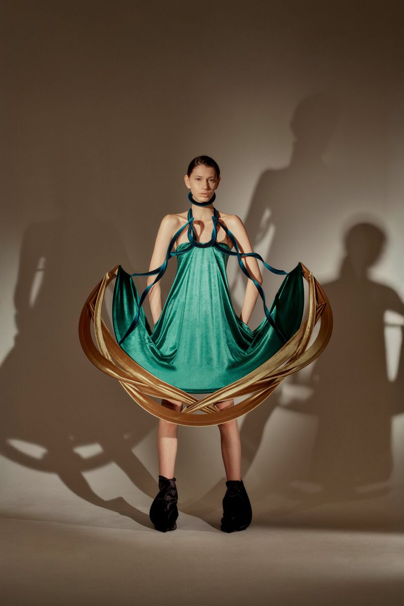 Fashionable woman in a stylish green dress with sculptural gold hoop detail, posing in a studio with dramatic shadow play.