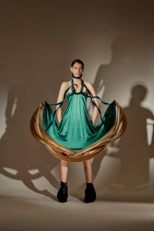 Fashionable woman in a stylish green dress with sculptural gold hoop detail, posing in a studio with dramatic shadow play.