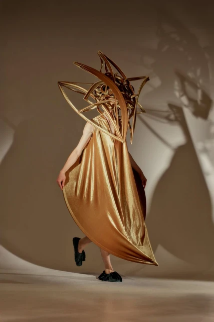 Person wears a gold metallic cape and sculptural gold headpiece in a studio setting. Fashion-forward, surreal design, creative concept.