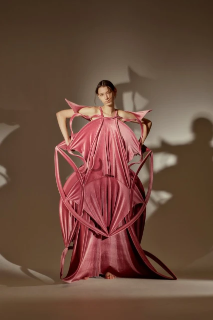 Young woman in an avant-garde, pink velvet gown poses in a studio with a neutral backdrop, her hands on her hips.