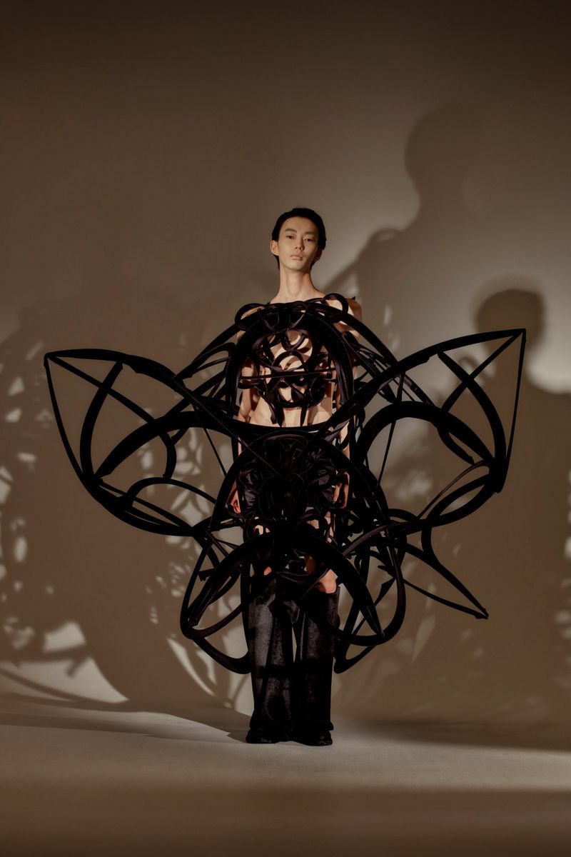 Model Gendai Funato wearing a large black sculptural wearable frame for the Ryunosukeokazaki S/S 26 lookbook in a lit studio.