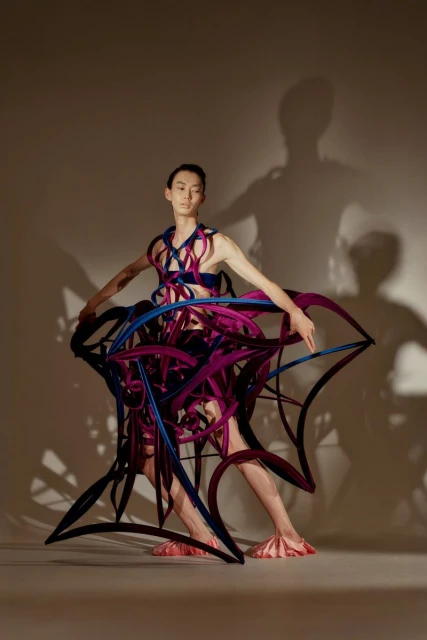 Eye-level shot of a single adult posing in a sculptural, colorful dress with a neutral background, creating a fashion-forward artistic statement.