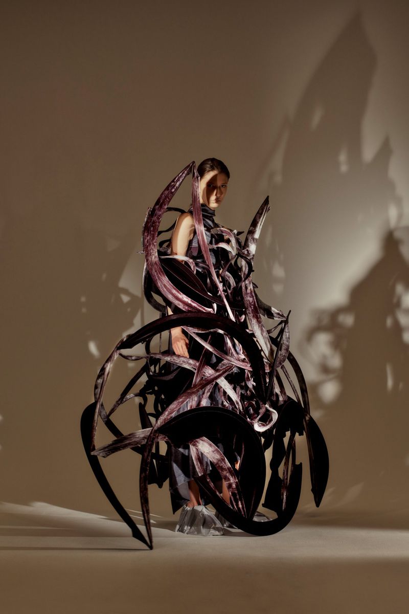 Woman poses in a surreal, dark, sculptural garment in a studio. Angular shadows stretch on the studio backdrop. Conceptual fashion.