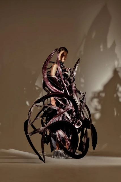 Woman poses in a surreal, dark, sculptural garment in a studio. Angular shadows stretch on the studio backdrop. Conceptual fashion.