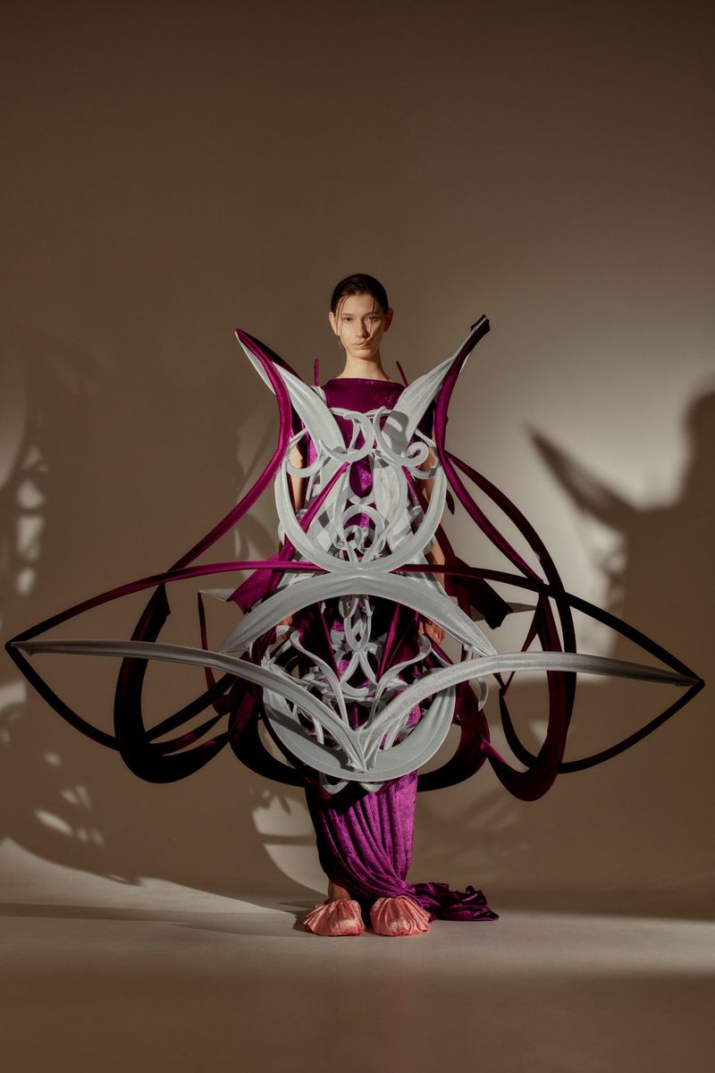 A fashion model poses in a studio wearing a sculptural purple dress with a white geometric design for a high-fashion editorial shoot.