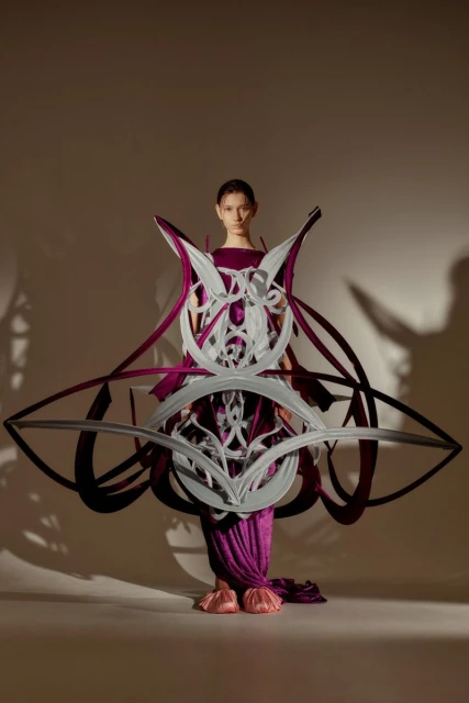 A fashion model poses in a studio wearing a sculptural purple dress with a white geometric design for a high-fashion editorial shoot.