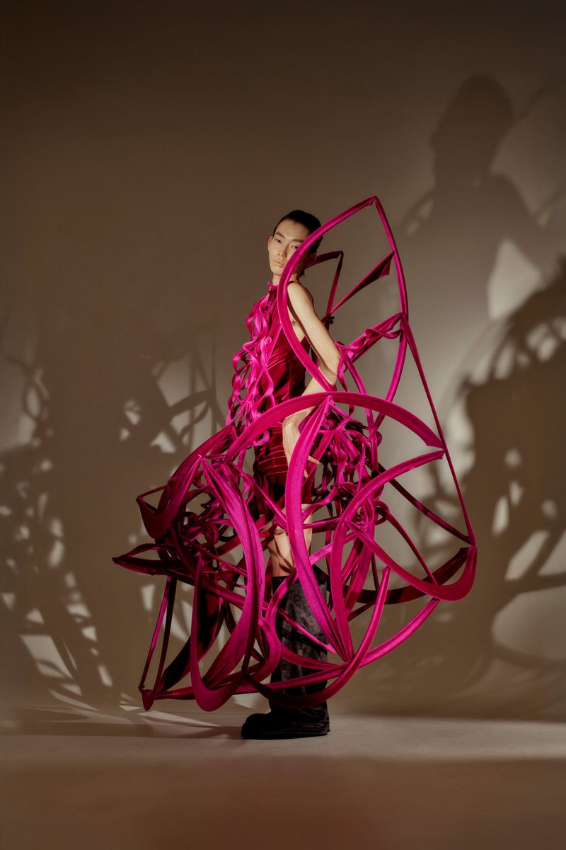 A striking editorial shot of a person in a pink sculptural dress, standing in a neutral-toned studio with creative lighting casting shadows.