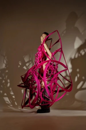 A striking editorial shot of a person in a pink sculptural dress, standing in a neutral-toned studio with creative lighting casting shadows.