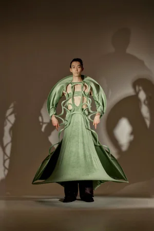 Model Gendai Funato wearing a sculptural sage green avant-garde garment with sweeping flared silhouette against a neutral wall with dramatic shadows.