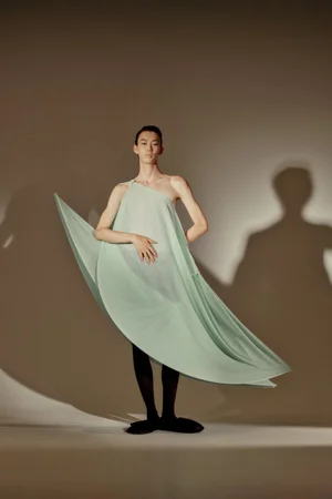 Model posing in an architectural mint green draped garment with black tights against a studio backdrop casting a sharp shadow.