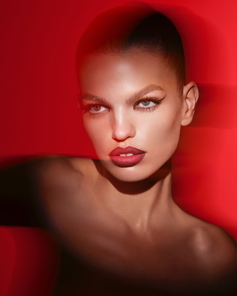 Close-up portrait of an adult woman with dramatic eye makeup, set against a vibrant red backdrop in a beauty fashion editorial.