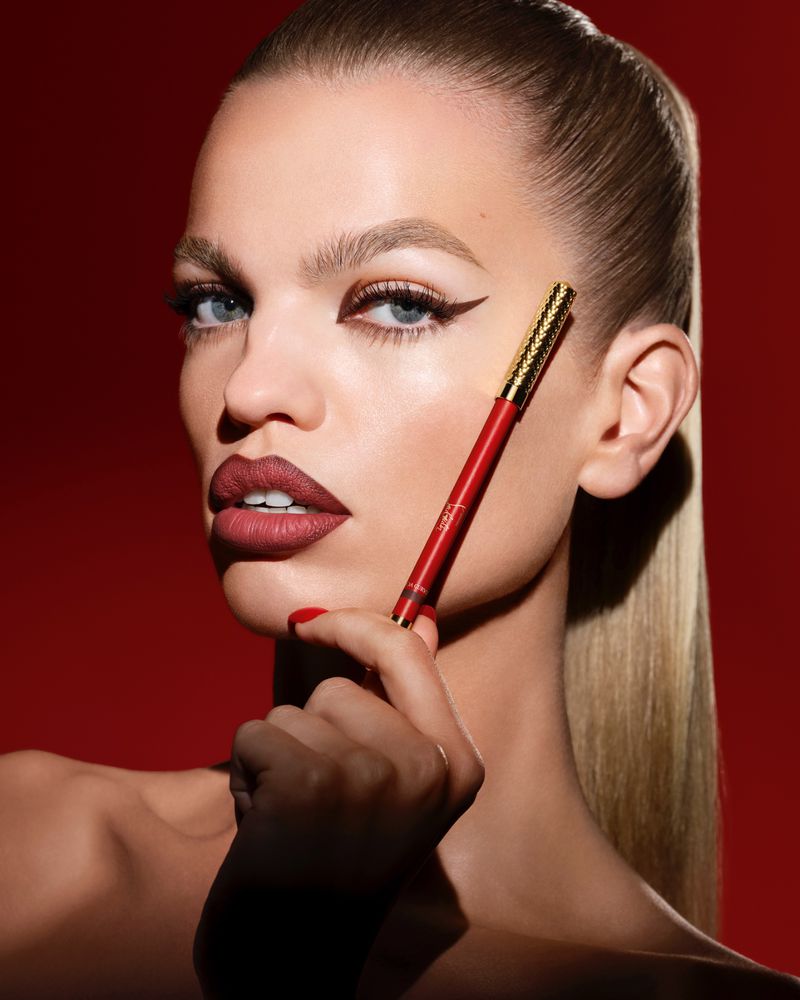 Close-up of a blonde woman with a red lip and winged eyeliner, holding a red and gold eyeliner pencil against a red background.