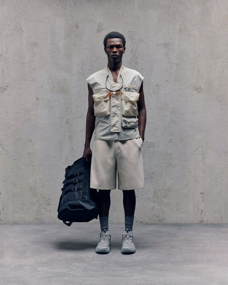 Male fashion model poses in studio wearing beige shorts and utility vest holding black backpack against a concrete wall backdrop.