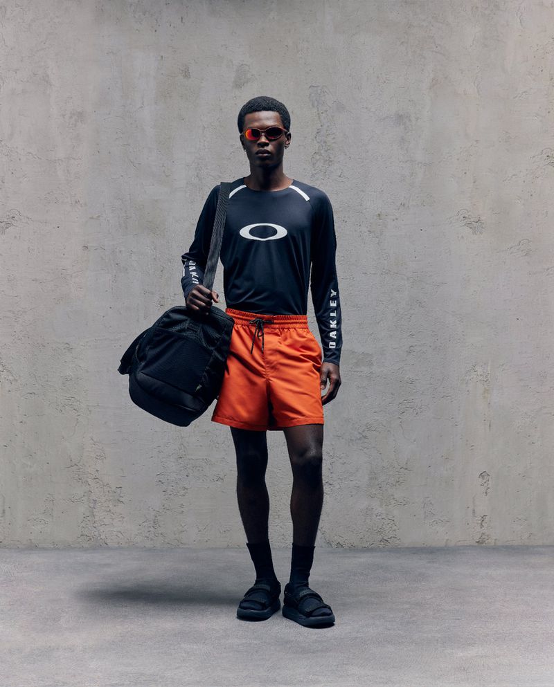 Athletic man in Oakley branded activewear, wearing sunglasses and carrying a black duffel bag, stands against a textured grey background.