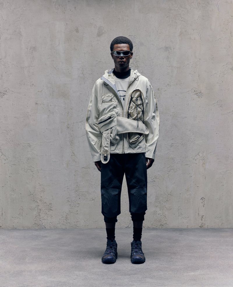 Man stands in grey jacket and black shorts against a textured grey wall, wearing sunglasses, with studio lighting and a minimalist aesthetic.