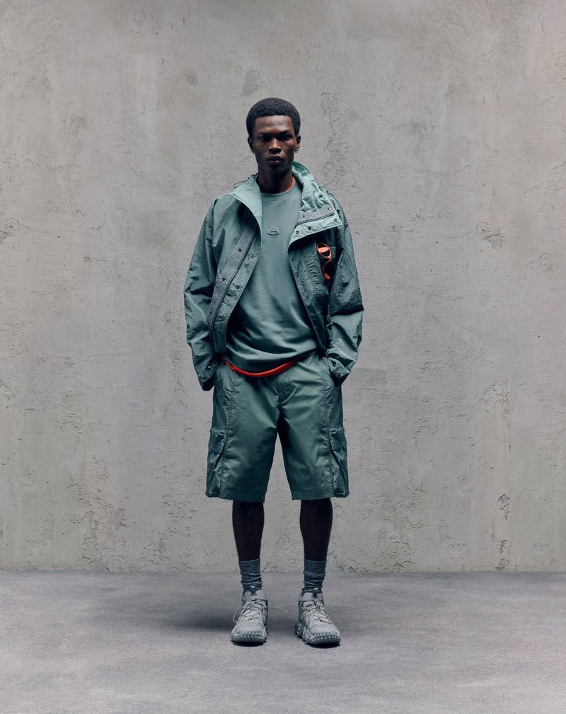 Full-length shot of a Black male model wearing an aqua-colored jacket, t-shirt, shorts and sneakers standing against a grey textured wall.