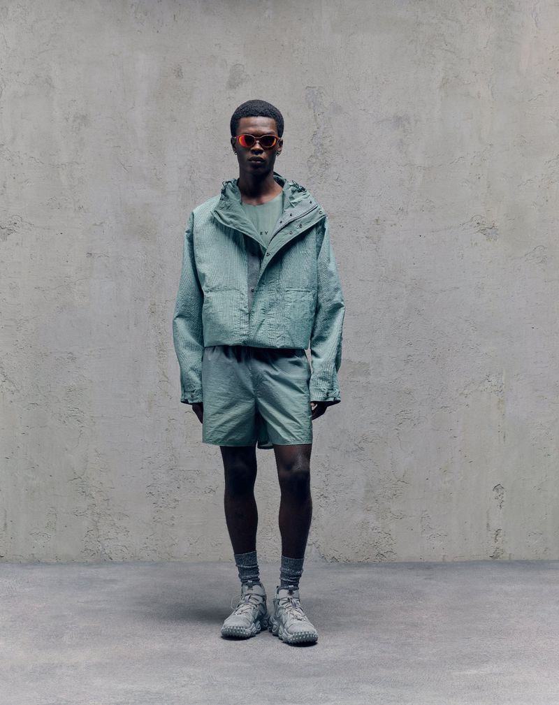 Man in mint green outfit with sunglasses, stands in a minimalist studio, showcasing fashion apparel against a neutral backdrop.
