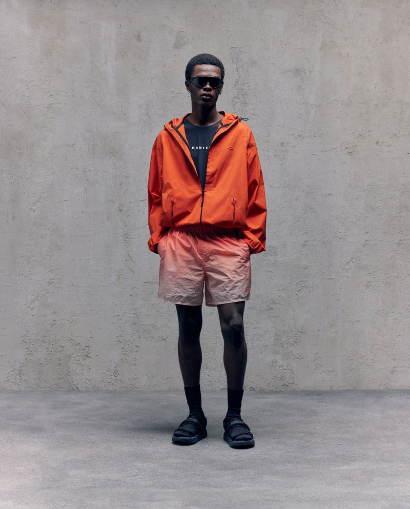 Man in orange hooded jacket and ombre shorts poses against textured gray wall. He wears sunglasses and sandals. Fashion shot. 