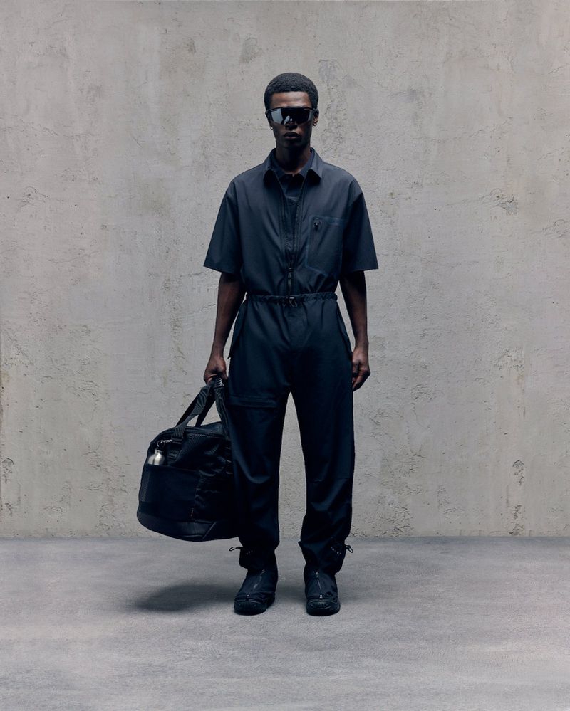 An adult male with dark skin poses in a black jumpsuit with a duffel bag against a textured grey wall in a studio setting.