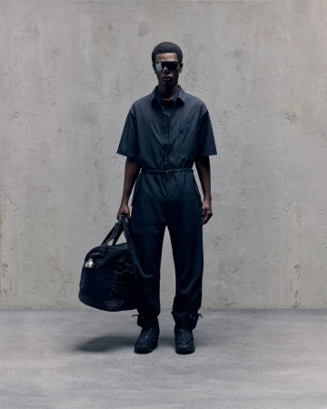An adult male with dark skin poses in a black jumpsuit with a duffel bag against a textured grey wall in a studio setting.