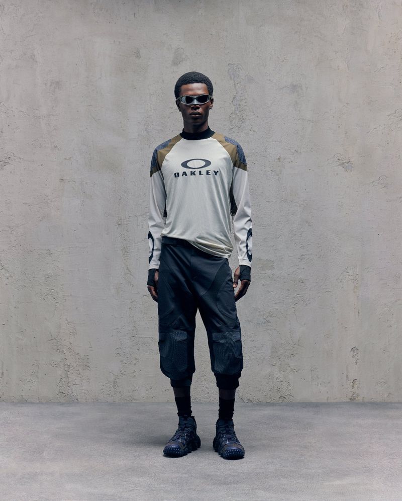 A slender Black male model strikes a pose indoors wearing an Oakley branded long-sleeved shirt, capri-length athletic pants, sunglasses, and sneakers.