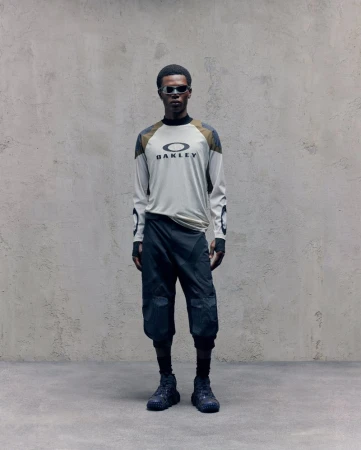 A slender Black male model strikes a pose indoors wearing an Oakley branded long-sleeved shirt, capri-length athletic pants, sunglasses, and sneakers.