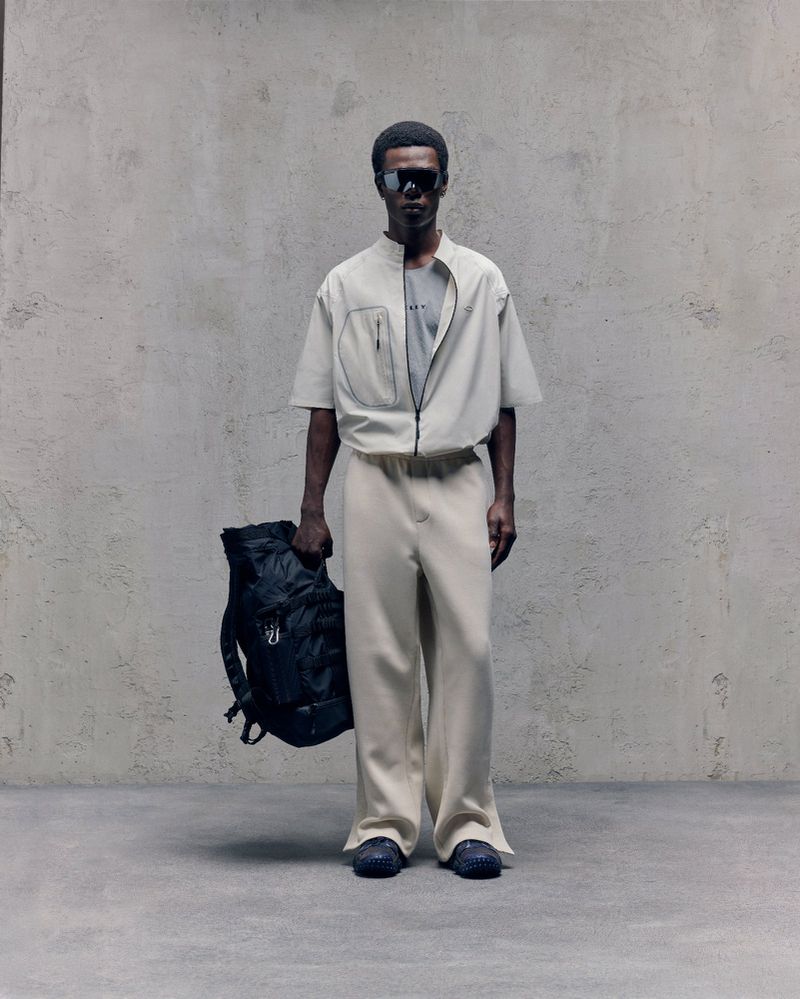 Man poses in neutral-toned zipper jacket and pants, holding a black backpack against a textured grey background, fashion forward.