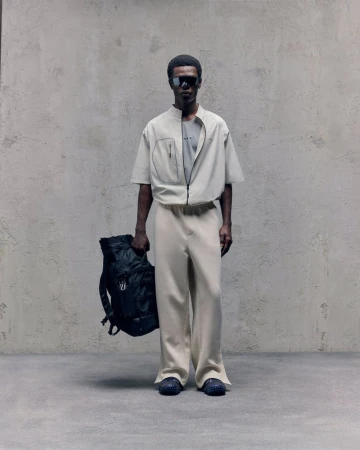 Man poses in neutral-toned zipper jacket and pants, holding a black backpack against a textured grey background, fashion forward.