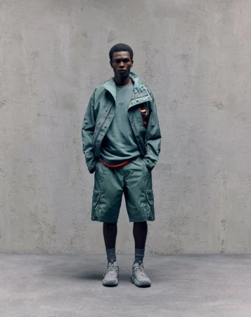 Model David Ikuepamitan wearing a green hooded jacket and matching cargo shorts against a textured grey wall for Oakley.