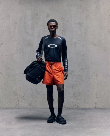 David Ikuepamitan wearing an Oakley long-sleeve shirt, orange shorts, and sunglasses against a textured studio backdrop.