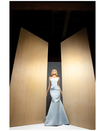 Woman in a light blue gown stands between two light wood panels in a studio with dark upper background and soft, diffused lighting.