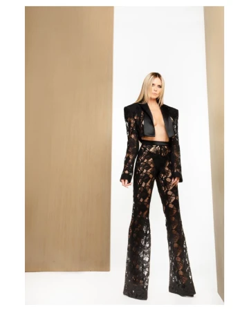 A woman poses in a black lace crop top and flared pants with a neutral-toned studio background, showcasing a fashion-forward look with dramatic flair.