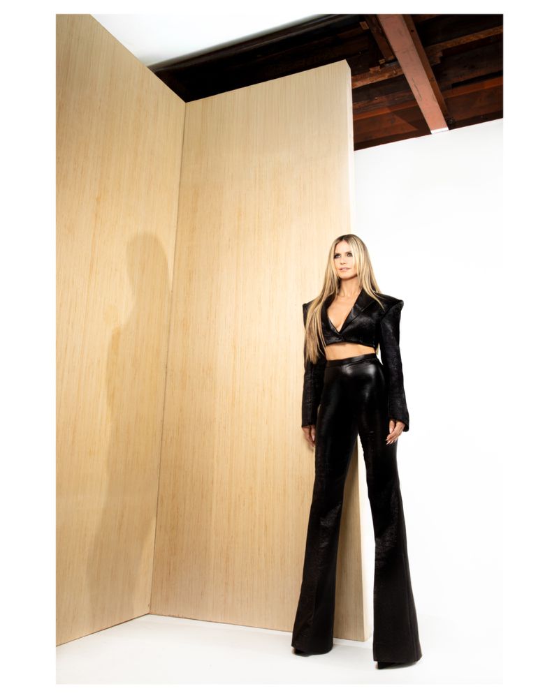 Blonde woman in a black two-piece outfit poses in a studio, standing near wood panels and a white wall under visible ceiling beams.