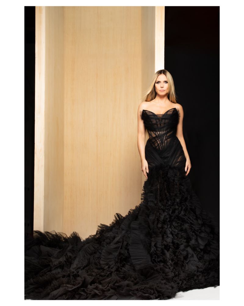 Heidi Klum poses in a strapless black gown with a voluminous ruffled train against a neutral background in a studio shot.