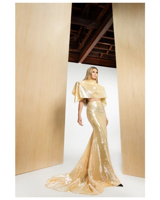 Adult female model poses in a shimmery gold two-piece dress with train in a bright indoor setting with light wood accents. 