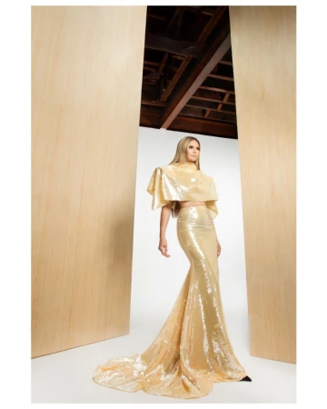 Adult female model poses in a shimmery gold two-piece dress with train in a bright indoor setting with light wood accents. 