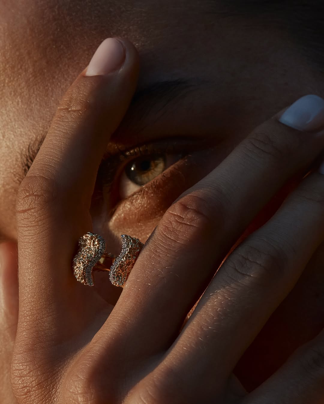 Close-up of a woman's face with a focus on the ring on her finger, showcasing beauty and jewelry details in soft lighting.