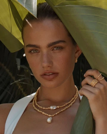 Close-up of an adult female model wearing gold jewelry, including a necklace, ring and hoop earrings, posed outdoors.