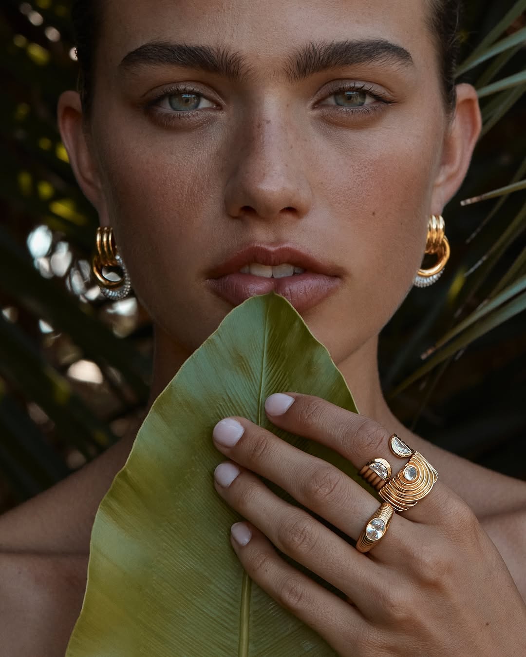 Close-up shot of a woman with tan skin and gold hoop earrings, holding a large green leaf in front of her mouth, with a neutral expression.