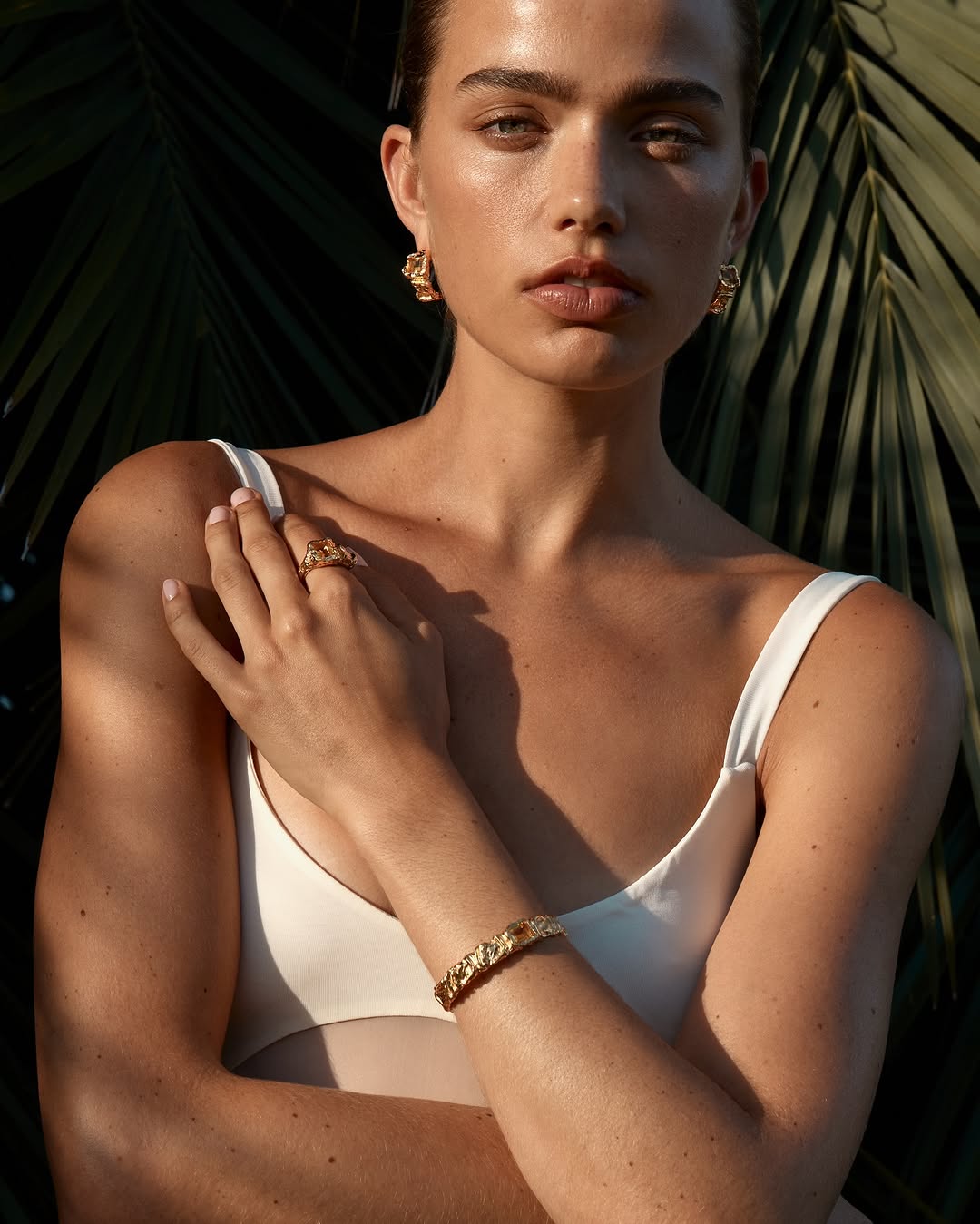 A young woman with fair skin poses outdoors wearing a white top and gold jewelry against a lush green backdrop of palm fronds.