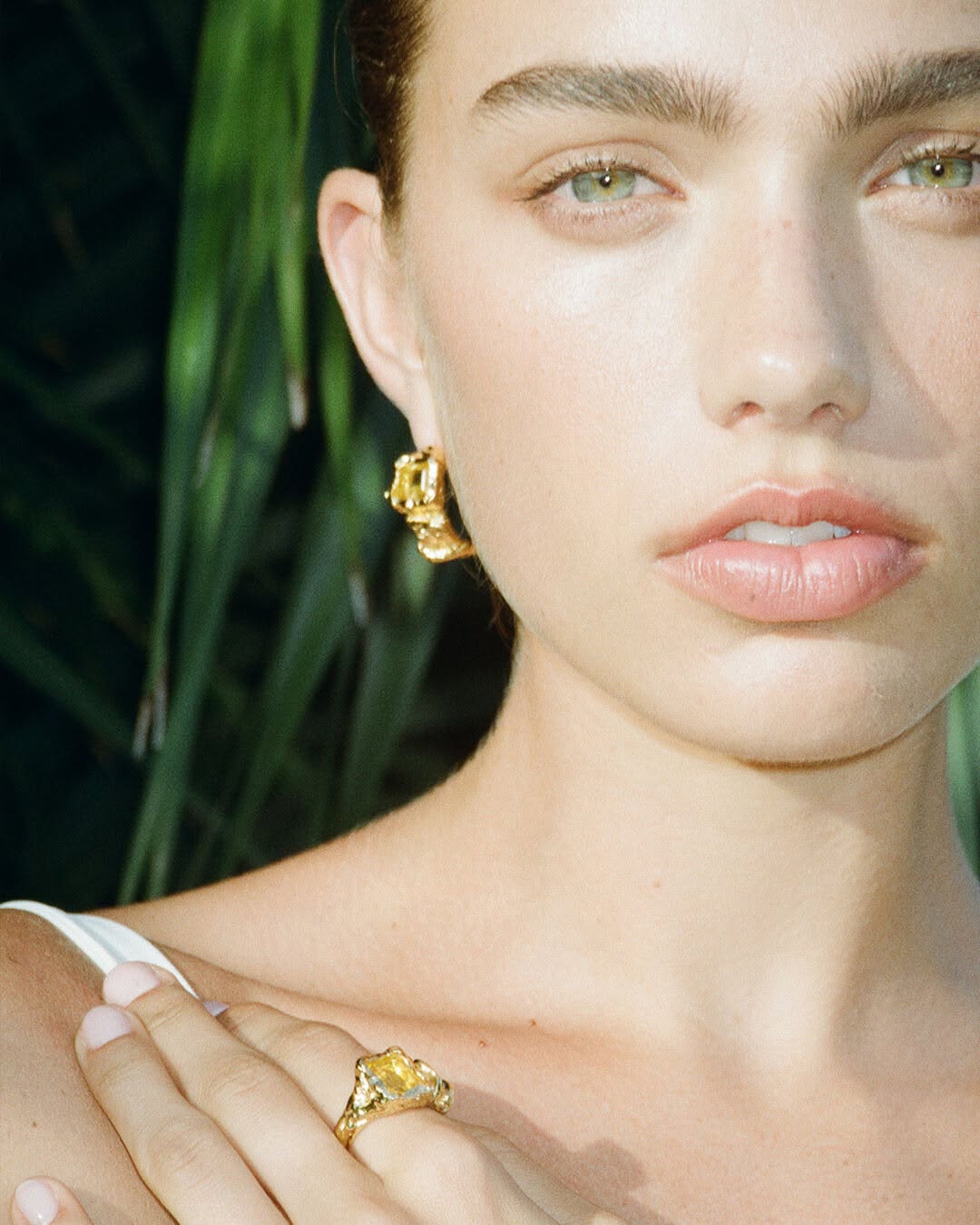 Close-up of a woman with gold hoop earrings and a ring, posing with green foliage in the background, jewellery advertising photography.