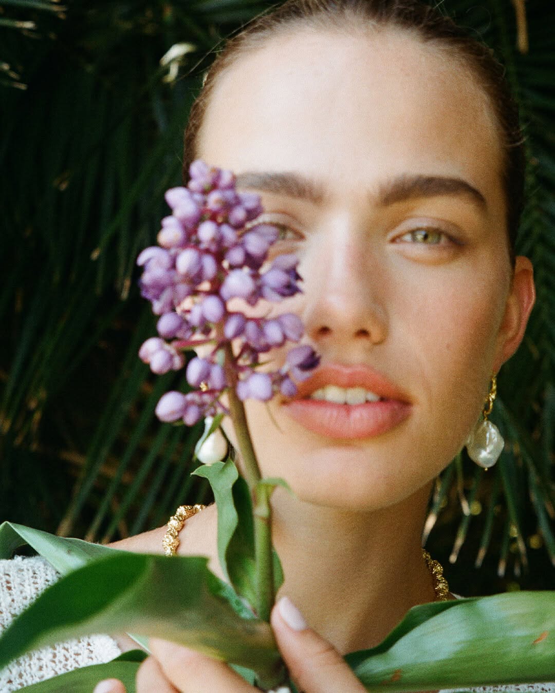 Close-up portrait of a fair-skinned young woman holding purple flowers in front of a green foliage background, showcasing beauty and nature.