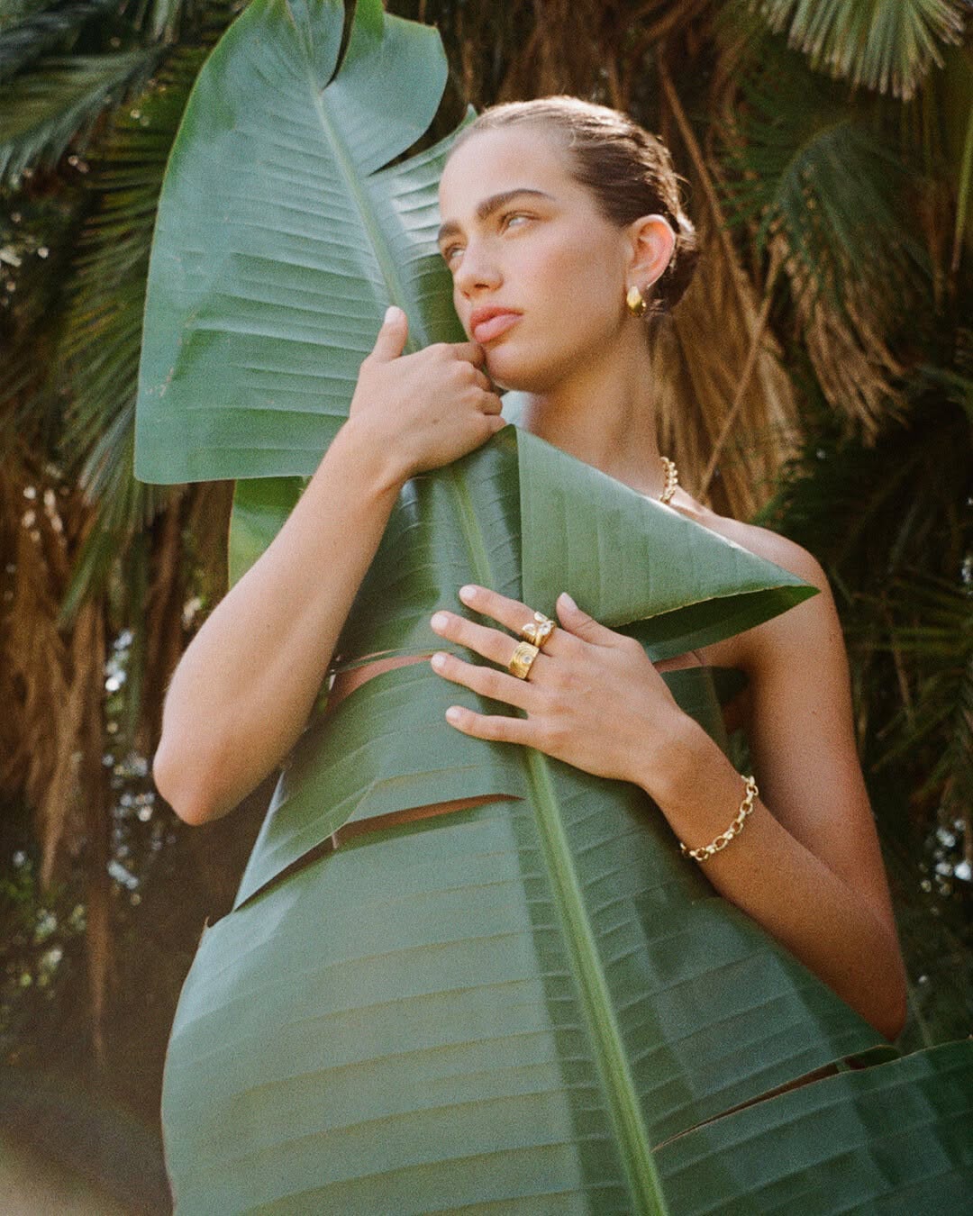 Portrait of woman surrounded by banana leaves in an outdoor setting, wearing gold jewelry including a necklace, bracelet, and ring.
