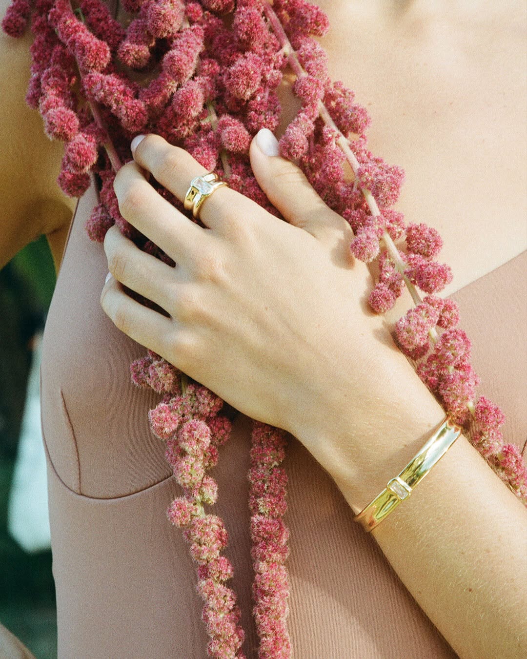 Close-up of a woman's arm and hand adorned with a gold bracelet and ring, posed with a pink floral stem.