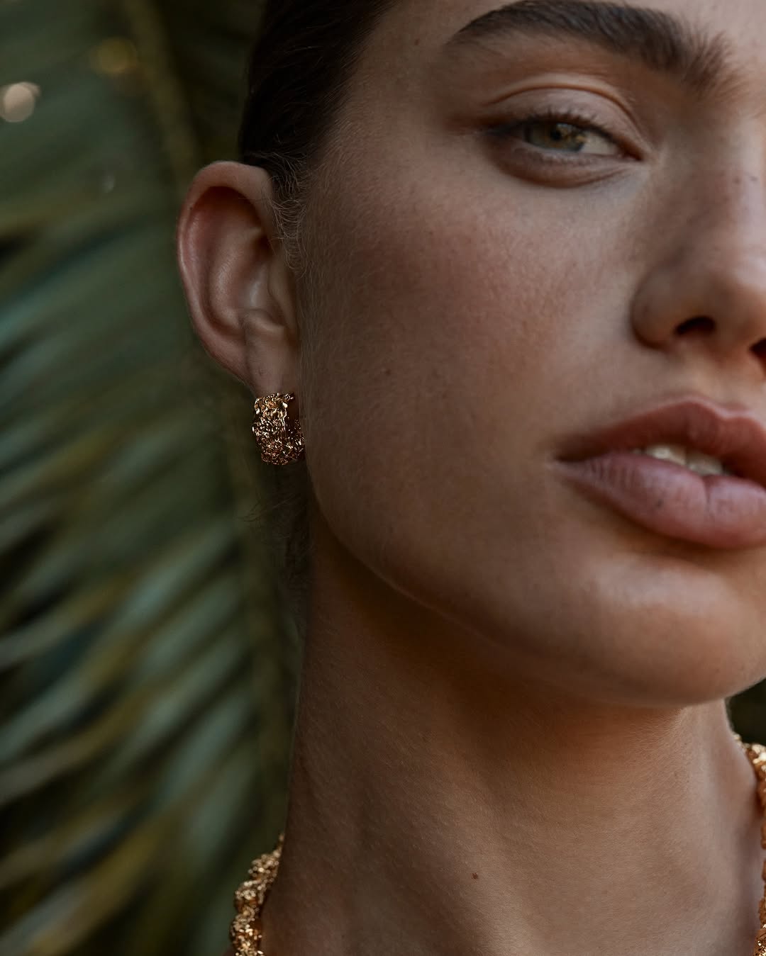 Close-up shot of an adult female model wearing gold jewelry including a necklace and earrings, with green foliage in the background.