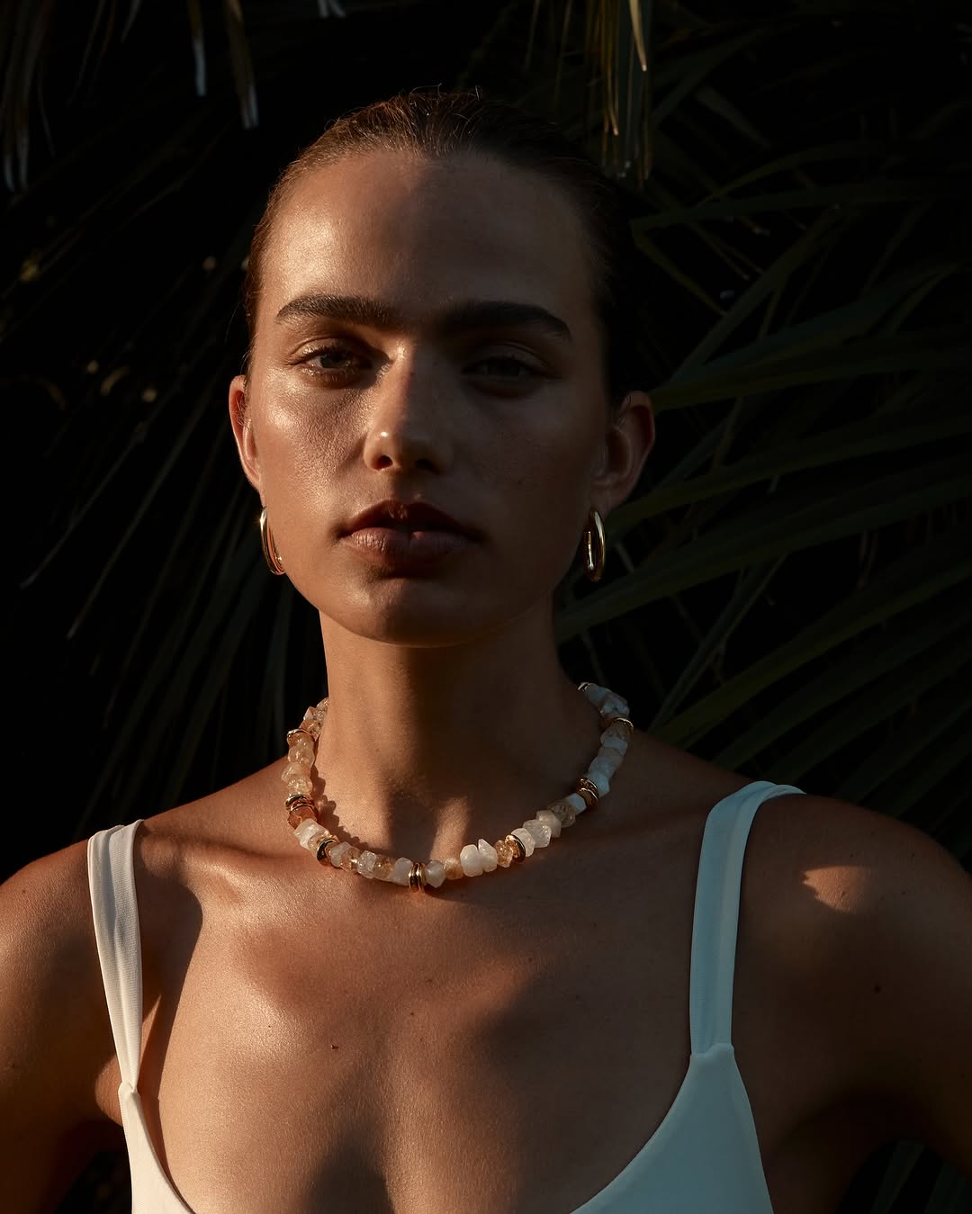 Close-up of a young woman wearing a beaded necklace and gold hoop earrings posing outdoors with palm leaves in the background.