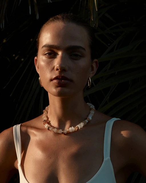 Close-up of a young woman wearing a beaded necklace and gold hoop earrings posing outdoors with palm leaves in the background.