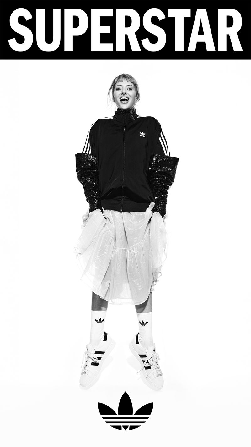 A female model in Adidas athletic wear poses in a black-and-white studio shot with the brand's logo and the word SUPERSTAR.
